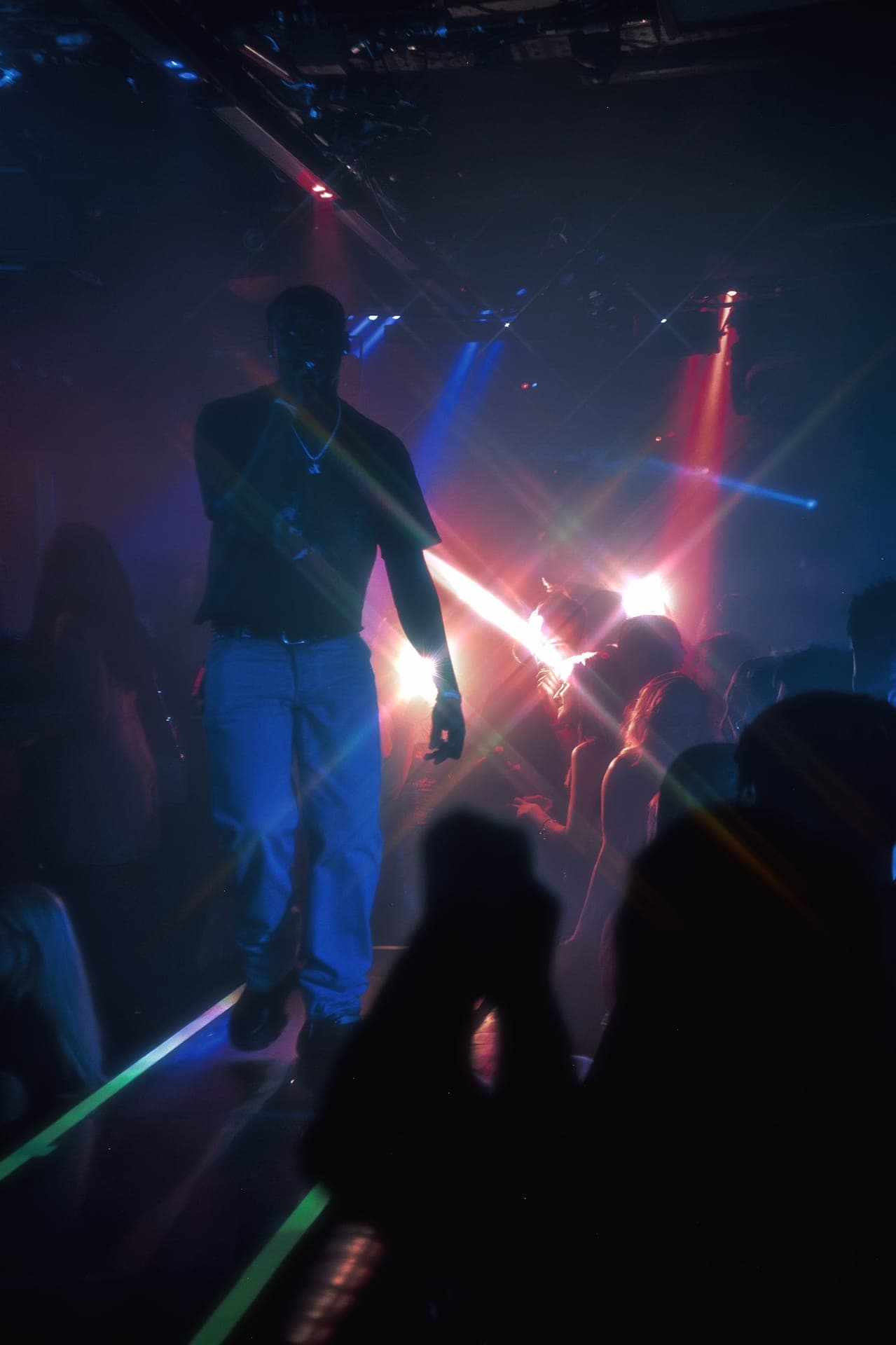 Silhouette of a performer on a lit nightclub stage above a dark crowd, with red and blue light beams
