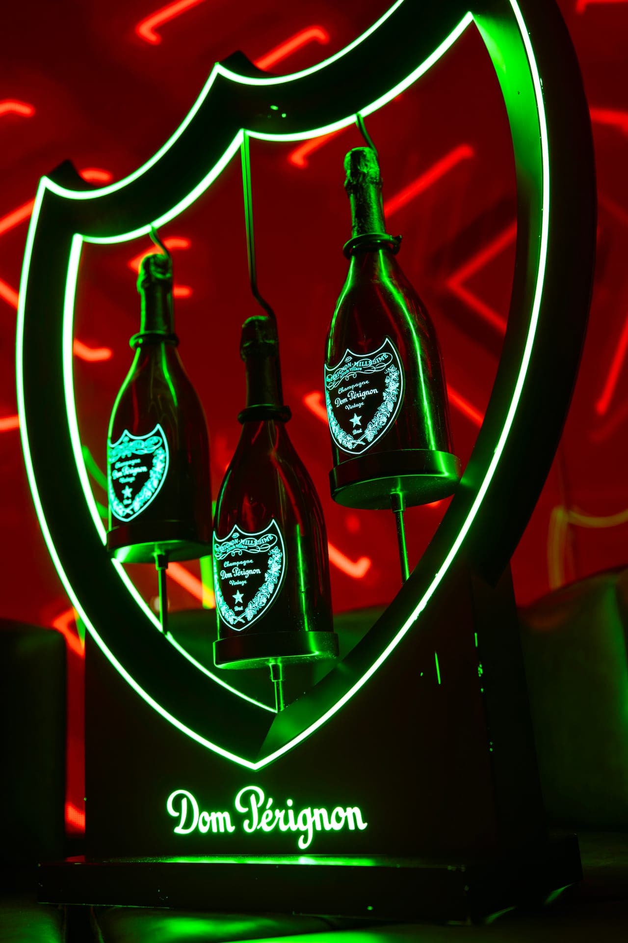 Three champagne bottles suspended in a lit green shield display against red neon