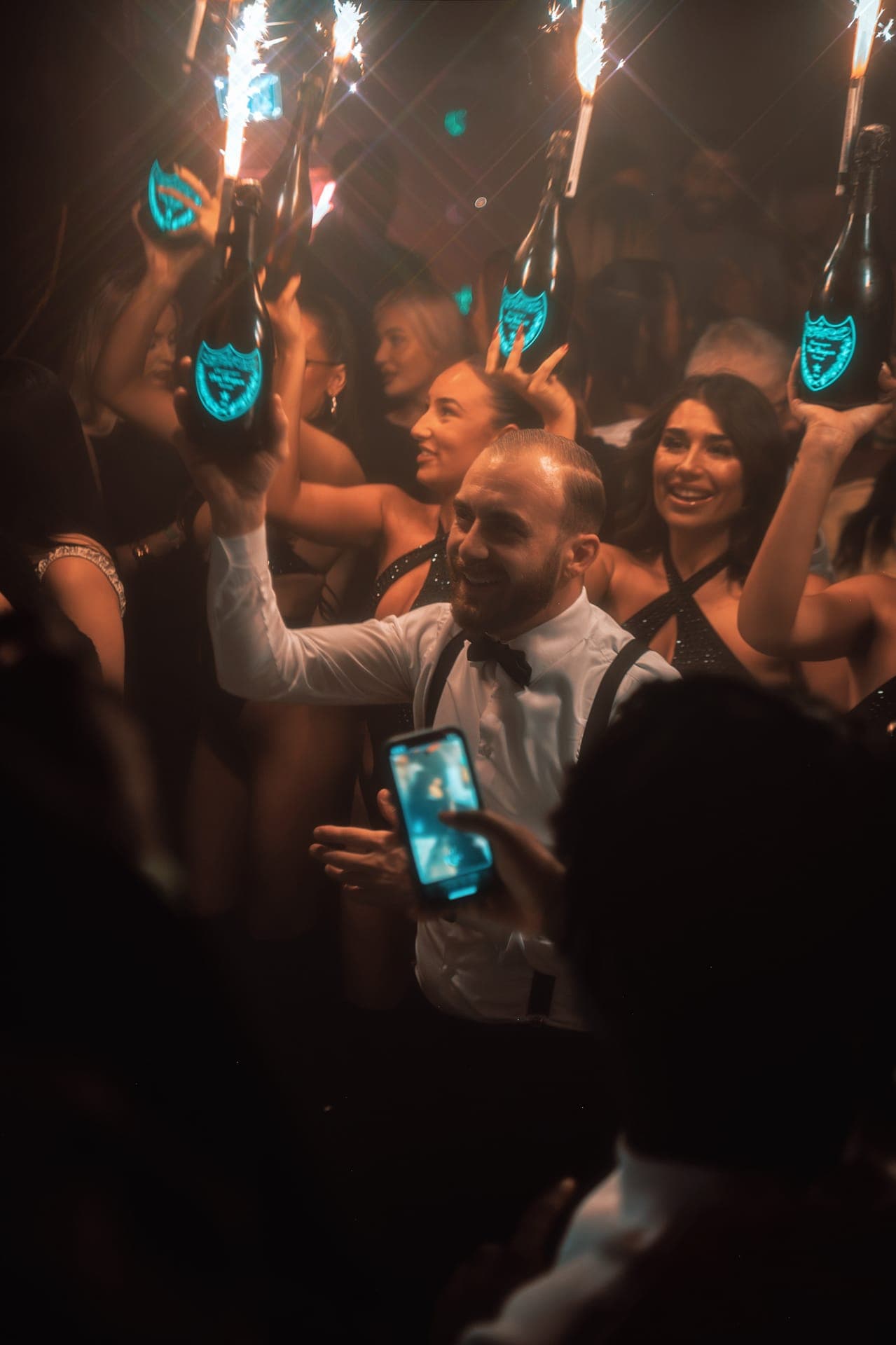 A man in a white shirt and bow tie celebrating among guests holding up sparkler-topped champagne bottles